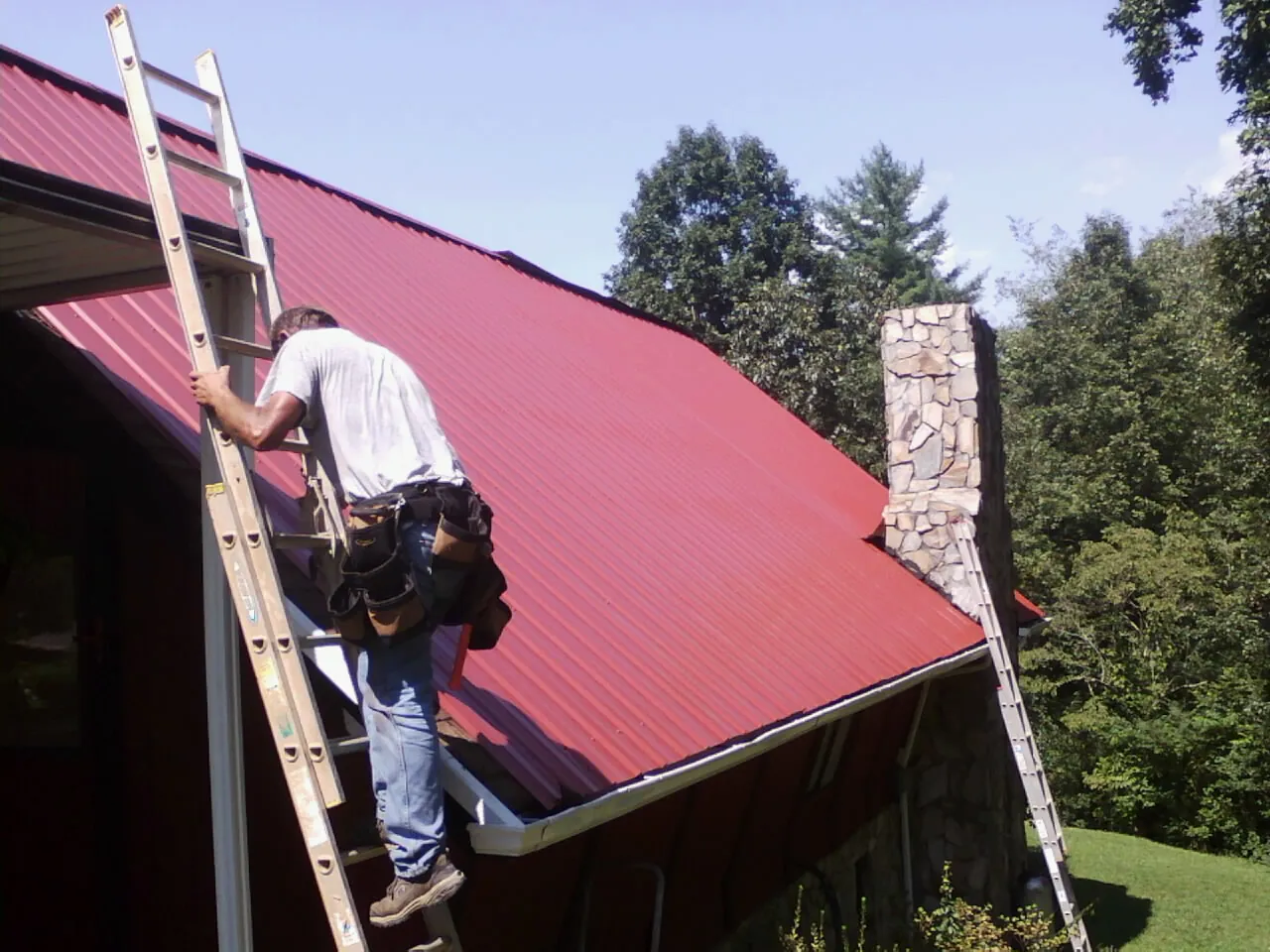 Roofer on a ladder beside a red metal roof and stone chimney — Soffit & Fascia Repair by Roofing Brothers in Pocahontas